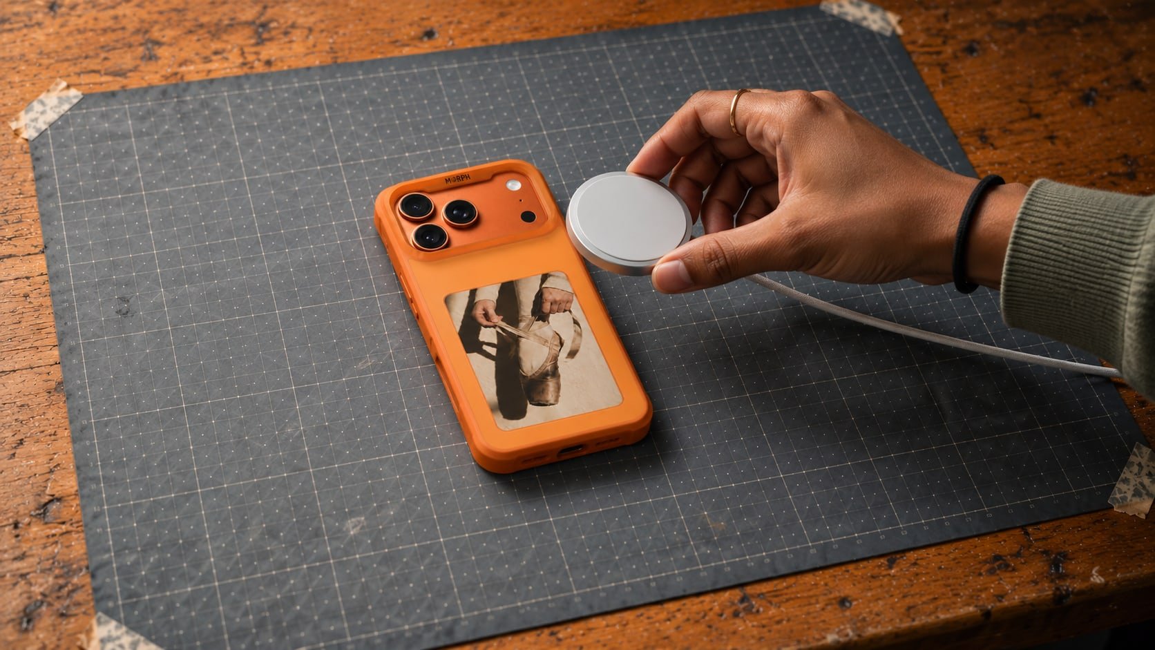 An orange Morph case lying panel up on a workbench mat, a photograph of ballet shoes on its e-ink screen, while a hand lowers a white MagSafe charger toward its back and stops just short of it.