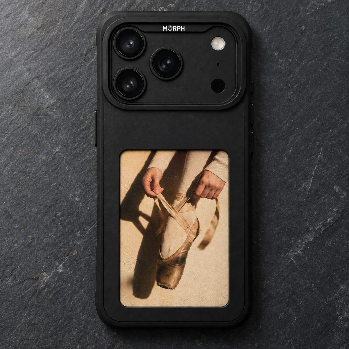 A black Morph Canvas Pro lying face up on dark slate, its panel showing a photograph of two hands tying the ribbons of a worn pointe shoe, the dancer up on the box in hard afternoon light.